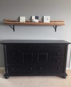 A big black dresser sits in front of a grey wall. There is a walnut wooden shelf above it. 