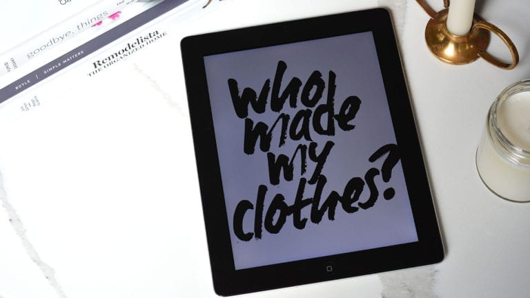 An iPad with the phrase "who made my clothes" is laid on a white surface and surrounded by a brass candle holder, a candle in a jar, and some books.