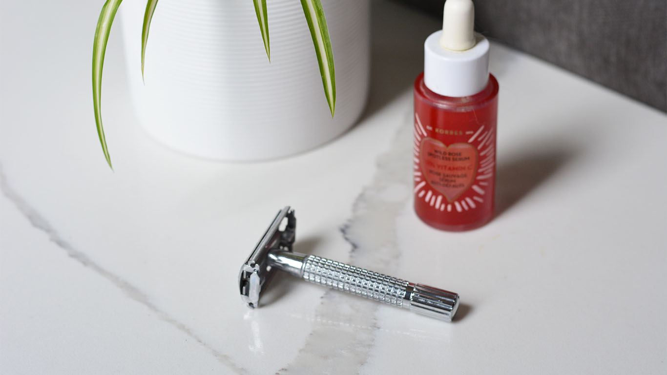 A sideview image of a zero waste stainless steel safety razor and a glass bottle of rose facial oil and part of a spider plant.