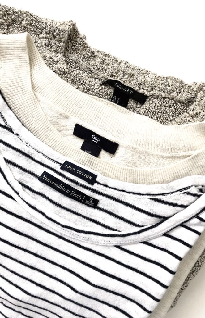 A slanted flat lay of a stack of 3 shirts from fast fashion brands, showing the labels. The top shirt is a black and white striped tee from Abercrombie & Fitch, the middle shirt is an oatmeal colored sweater from Gap, and the Botton shirt is a great sweater from Forever 21. All tops are thrifted.