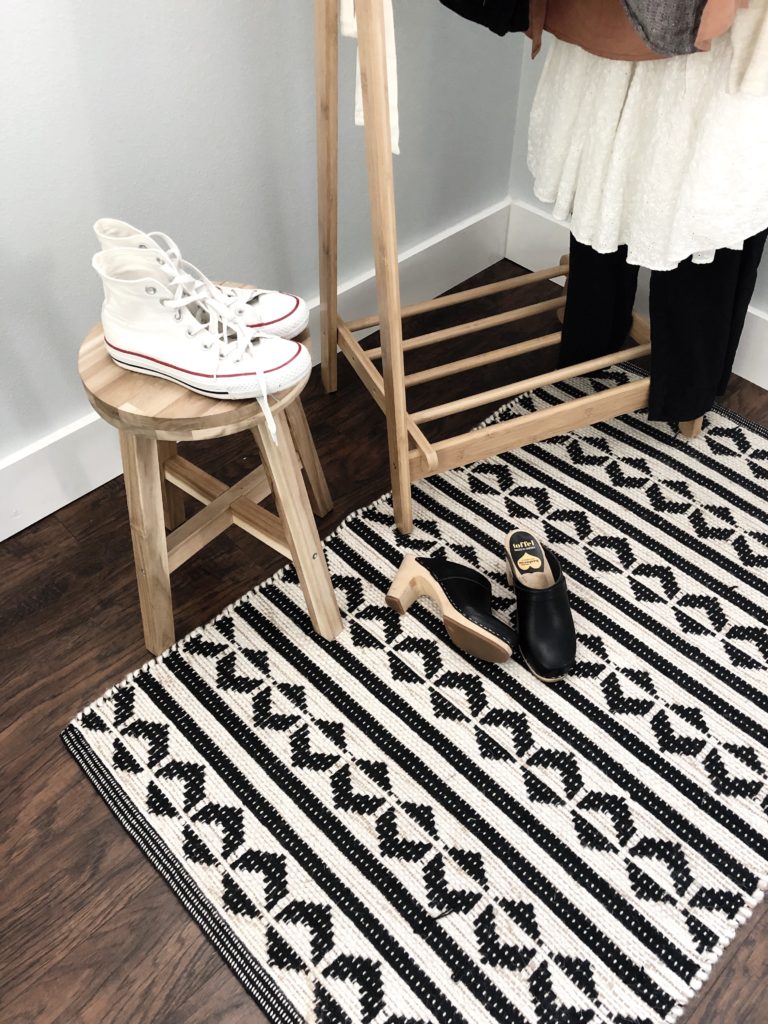 A shot taken from above of a pair of black clog shoes on a black and white rug and a pair of white high top converse on a light wood stool. The clothing rack is partially in the background.