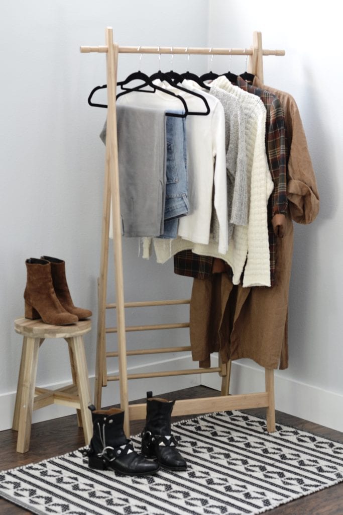 A clothing rack displaying clothing items for a cold weather capsule wardrobe. From left to right, there is a pair of grey wool pants, a pair of light blue jeans, a cream mock neck long sleeve top, a grey sweater, a cream colored sweater, a plaid blazer and a caramel robe. A pair of black boots sit on the floor and a pair of brown suede boots are on a wood stool next to the clothing rack.