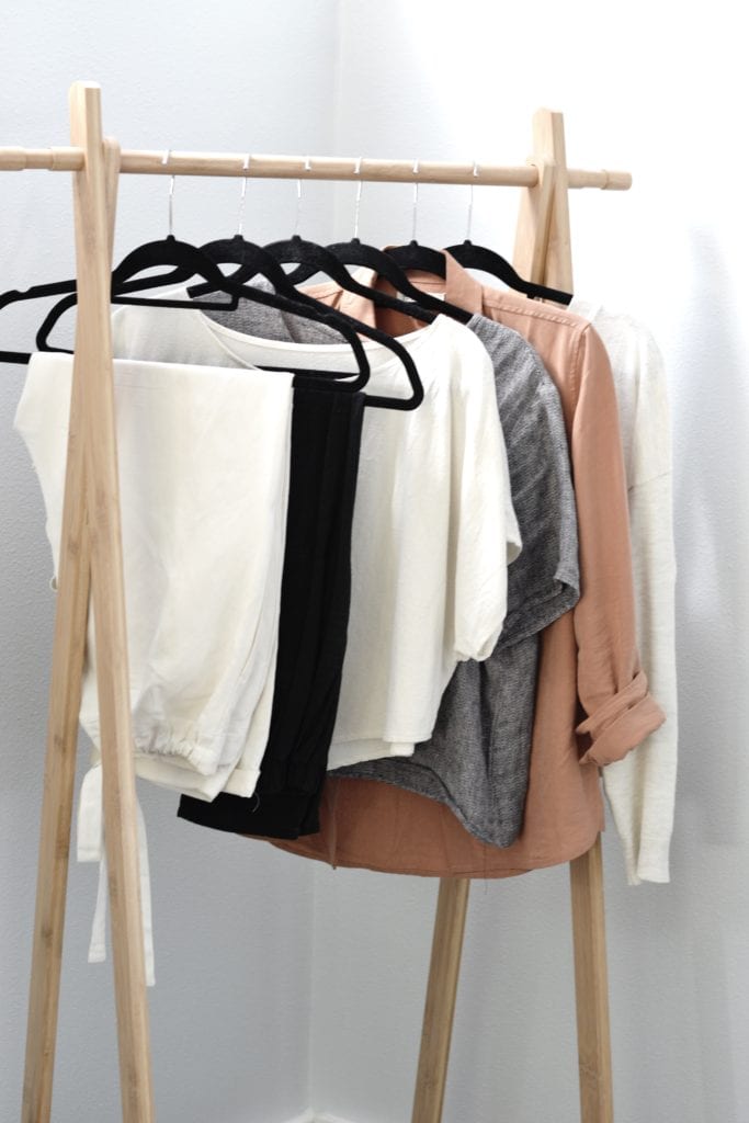 A light wood clothing rack holds six clothing items. From left to right, white pants, black pants, a white short sleeve top, a grey short sleeve top, an orange button up shirt and an oatmeal colored sweater.