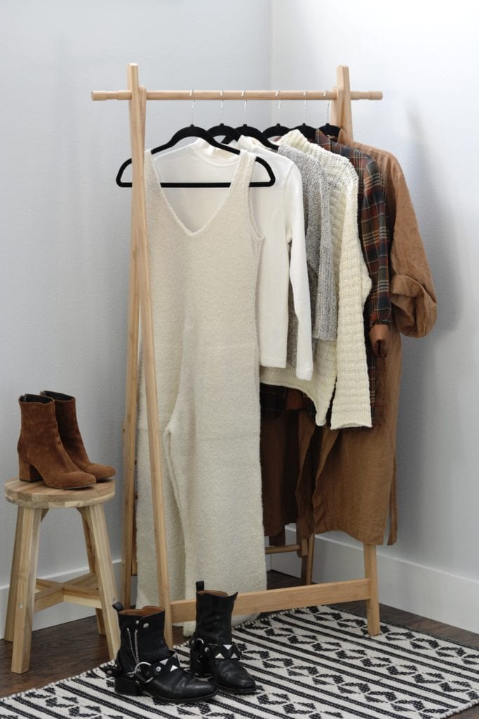 A clothing rack displaying clothing items for a cold weather capsule wardrobe. From left to right, there is a cream wool v-neck jumpsuit, a cream mock neck long sleeve top, a grey sweater, a cream colored sweater, a plaid blazer and a caramel robe. A pair of black boots sit on the floor and a pair of brown suede boots are on a wood stool next to the clothing rack.
