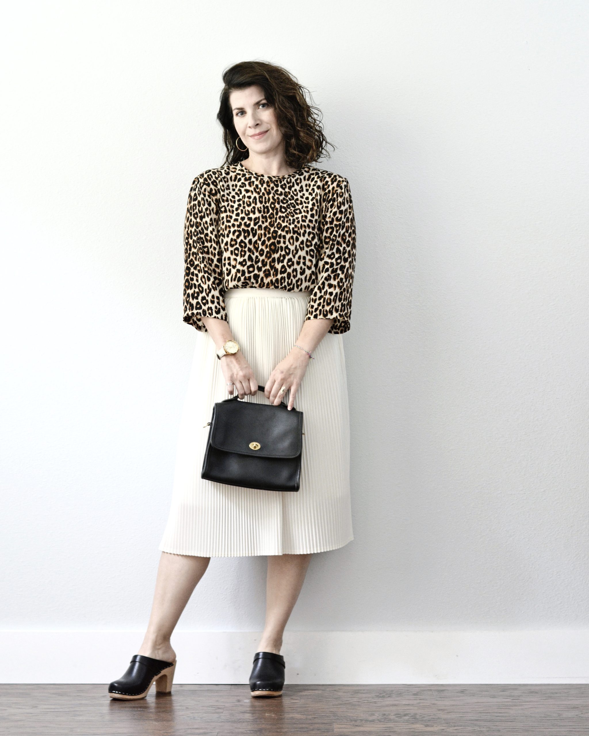 A woman is standing in front of a light grey wall. She is wearing a leopard print dress tucked into a white pleated skirt. She is holding a black handbag with both hands and she is wearing black clog shoes.