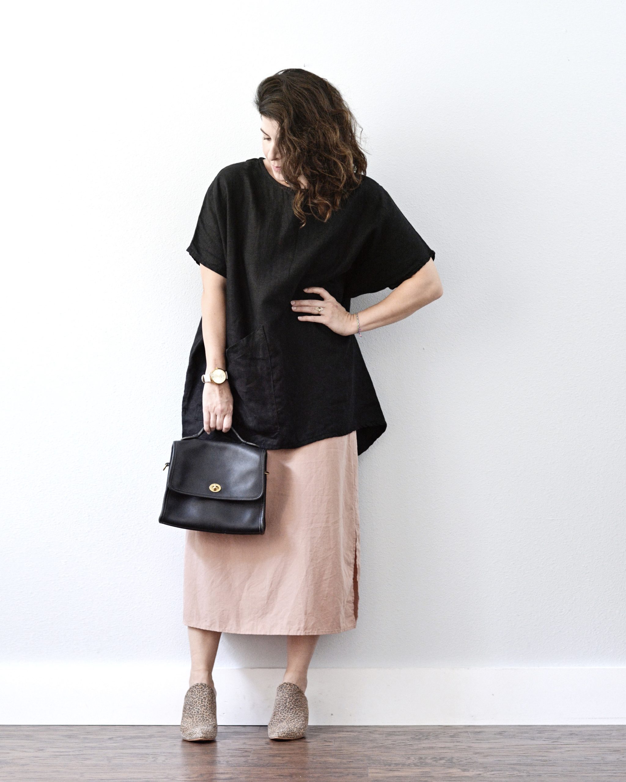 A white woman is standing against a light grey wall. She is wearing a black shirt sleeve tunic over a light pink dress. She is wearing leopard print mules and holding a black handbag in the right hand. Her left hand is on her left hip.