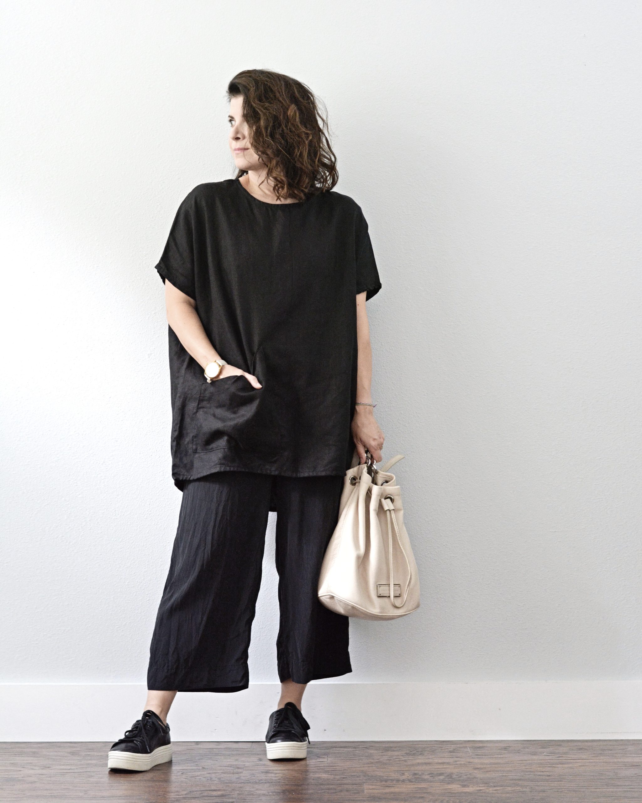 A white woman is standing against a light grey wall. She is wearing a black tunic top over a black wide leg jumpsuit so the jumpsuit looks like pants. She is wearing black sneakers with a white platform and holding a white handbag with her left hand. Her right hand is in her tunic pocket.