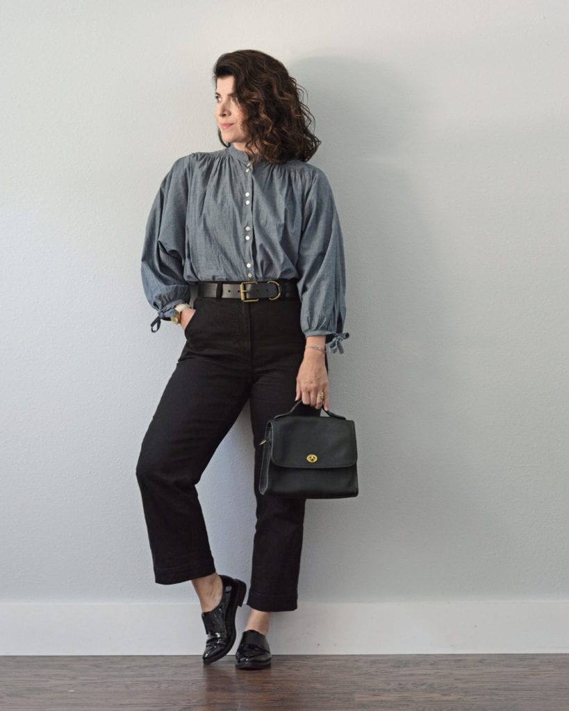 A woman is standing against a light grey wall. She is wearing a medium blue chambray shirt tucked into a pair of black pants with a black belt around the waist. She is holding a black handbag and is wearing black patent oxfords. Her right hand is in her pocket and her right leg is bend slightly. Her brown hair is left down and it is curly.