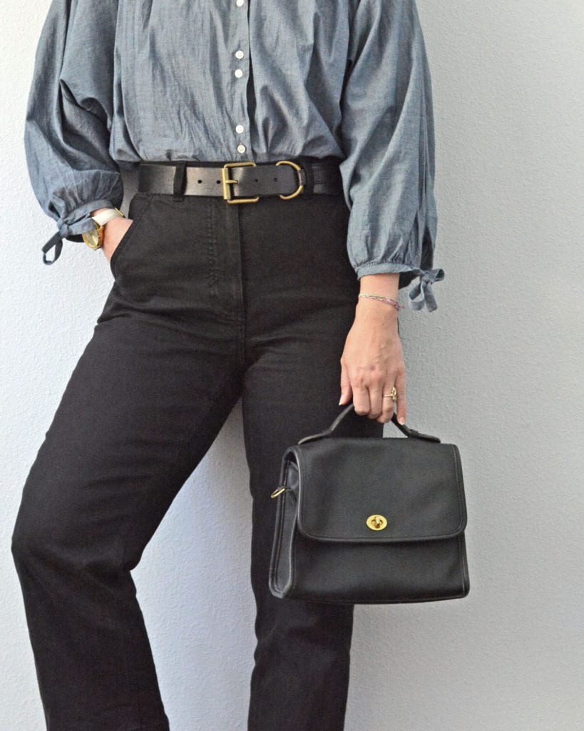 A closeup of a woman's torso and top part of her legs standing against a light grey wall. You can see her pants, handbag and watch more closely.