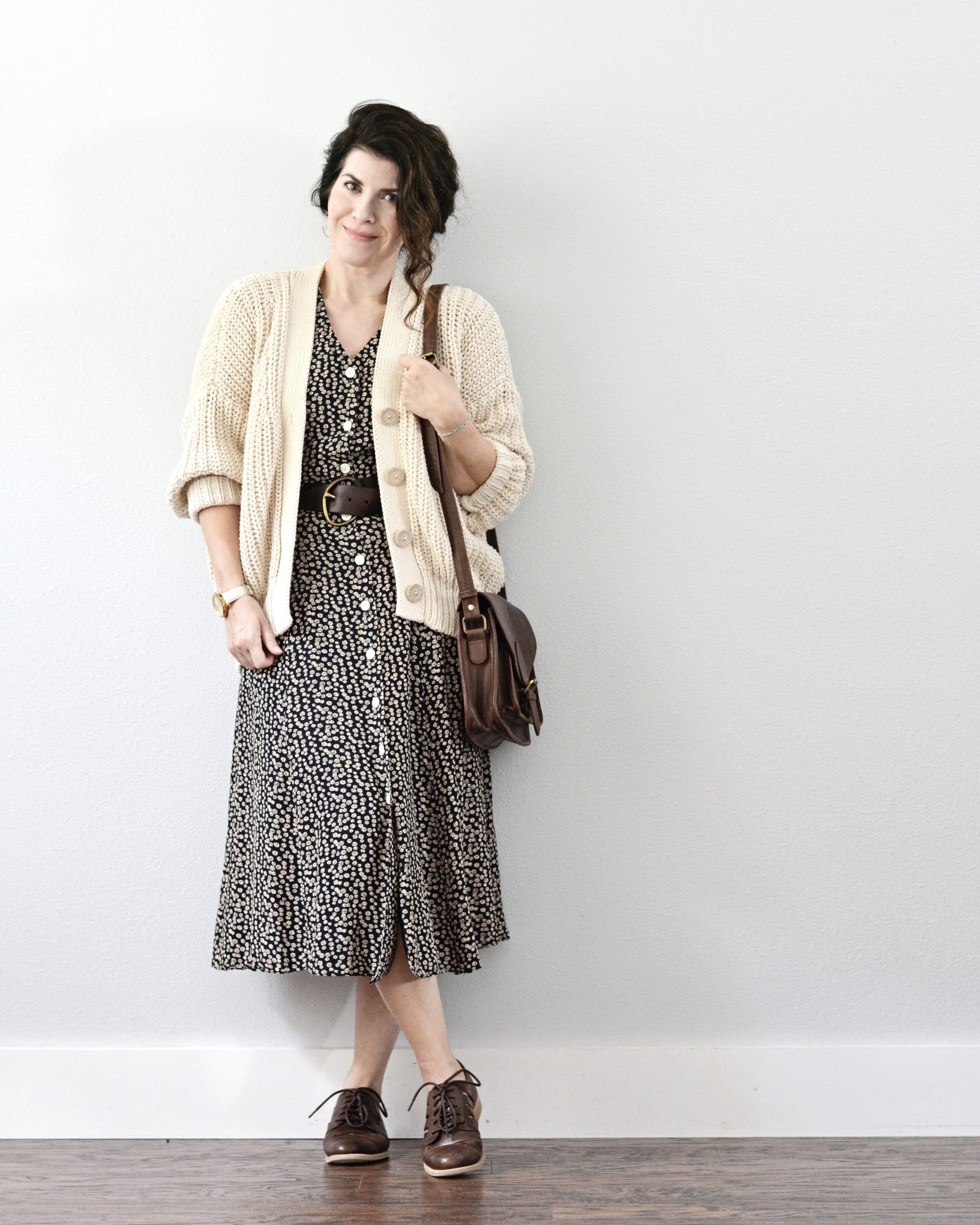 A woman standing against a light grey wall is wearing a navy floral dress with small tan flowers and a cream cardigan. You can see part of a brown belt where the cardigan opens and she is wearing brown Oxford shoes and a brown handbag hangs from her left shoulder. Her brown hair is pinned back.
