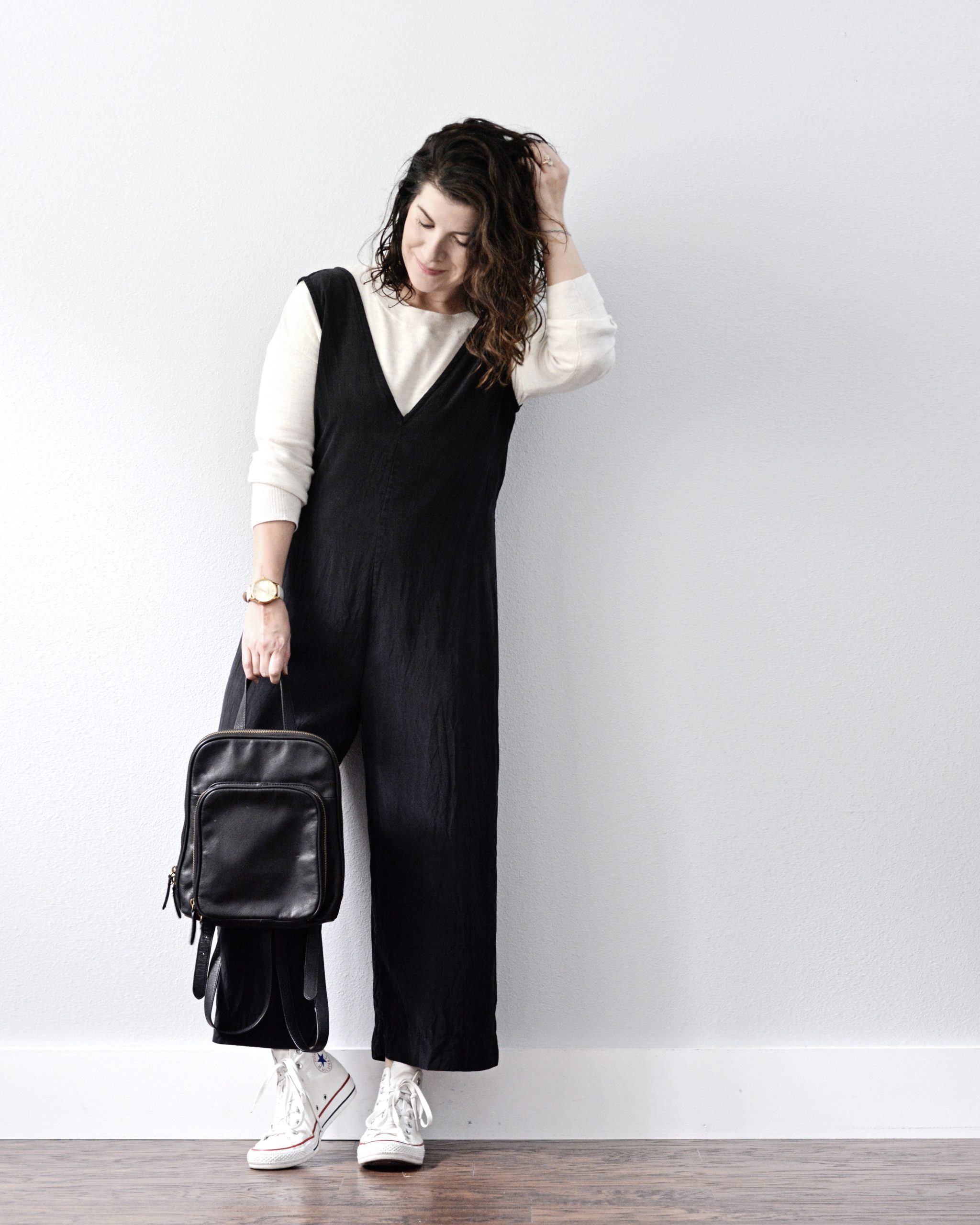 A white woman standing against a light grey wall is wearing a black v neck jumpsuit over an oatmeal colored sweater with white converse sneakers. She is holding a black leather backpack in her right hand and her left hand is in her hair. She is looking down.