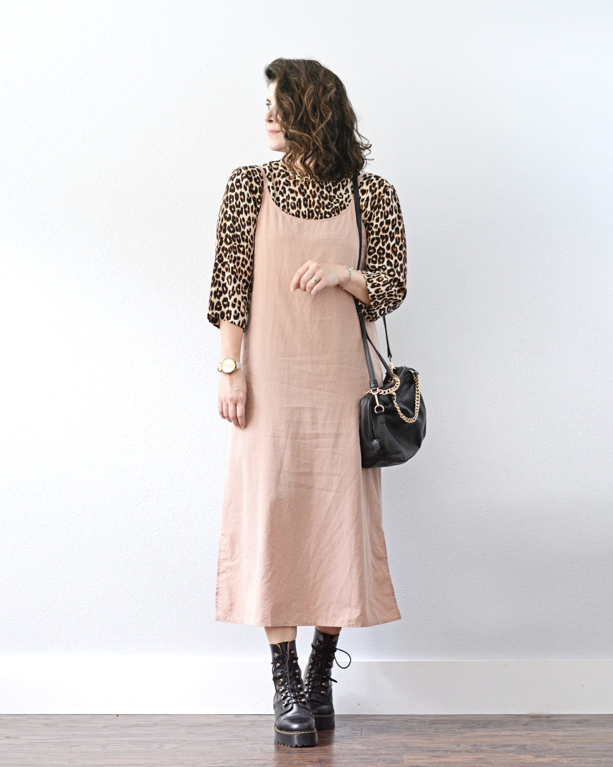 A woman standing against a light grey wall is looking to the right. She is wearing a light pink slip dress over a leopard print three quarter sleeve dress with black combat boots and a black handbag that is hanging from her left shoulder.