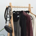 A partial view of a rack of clothing to be styled for this Pinterest inspired outfit post. The rack contains a grey dress, a black top, a Burgundy leather jacket, a white mock neck top and a camel wool coat. There is also a small black handbag and a black belt hanging at the front.