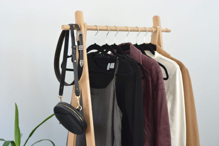 A partial view of a rack of clothing to be styled for this Pinterest inspired outfit post. The rack contains a grey dress, a black top, a Burgundy leather jacket, a white mock neck top and a camel wool coat. There is also a small black handbag and a black belt hanging at the front.