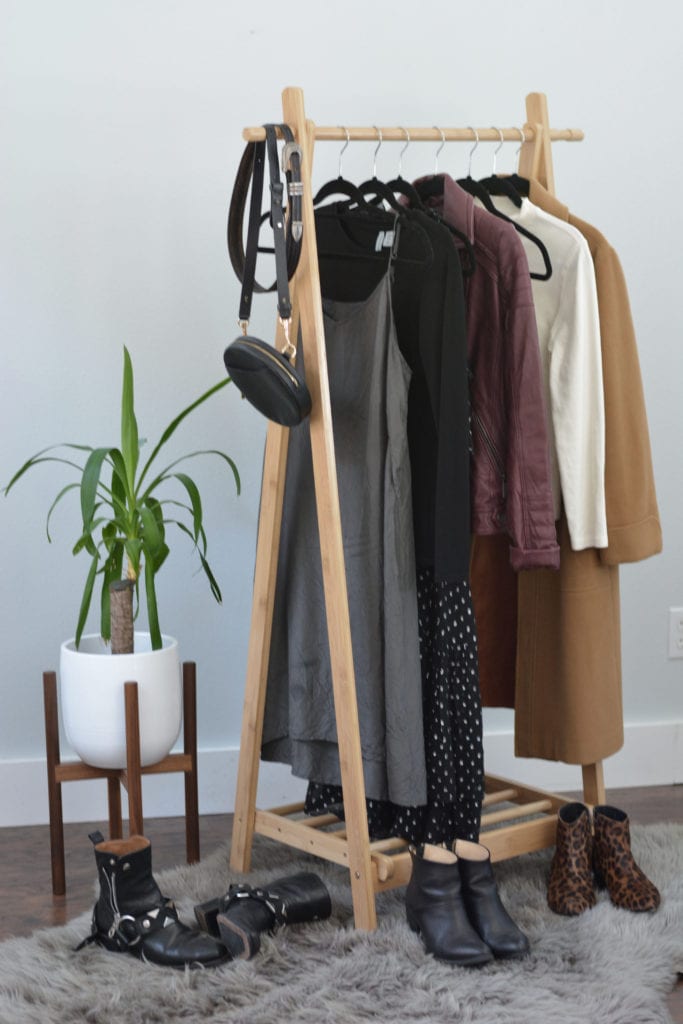 A rack of clothing to be styled for this Pinterest inspired outfit post. The rack contains a grey dress, a black top, a burgundy leather jacket, a white mock neck top and a camel wool coat. There is also a small black handbag and a black belt hanging at the front. A green yucca tree plant is to the left of the rack and 3 pairs of shoes are at the base, scattered over a great fur rug.