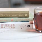 A stack of 3 books laid flat with spines showing - "The Millionaire Next Door", A Year of Less" and "Mending Matters" next to a glass of coffee.