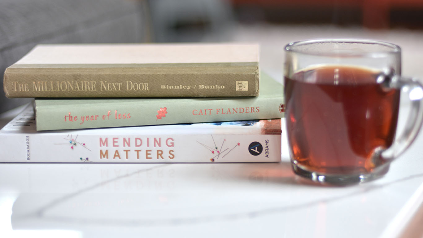 A stack of 3 books laid flat with spines showing - "The Millionaire Next Door", A Year of Less" and "Mending Matters" next to a glass of coffee.