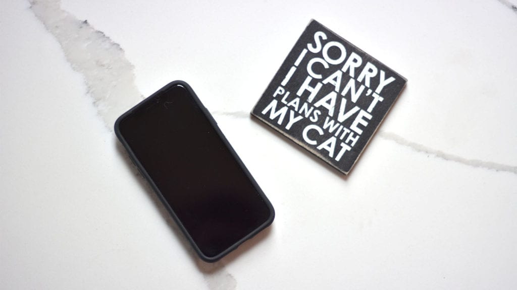 An overhead shot of my iPhone with a black screen and a coaster that reads "Sorry I can't I have plans with my cat". Stop shopping by removing apps on your phone.