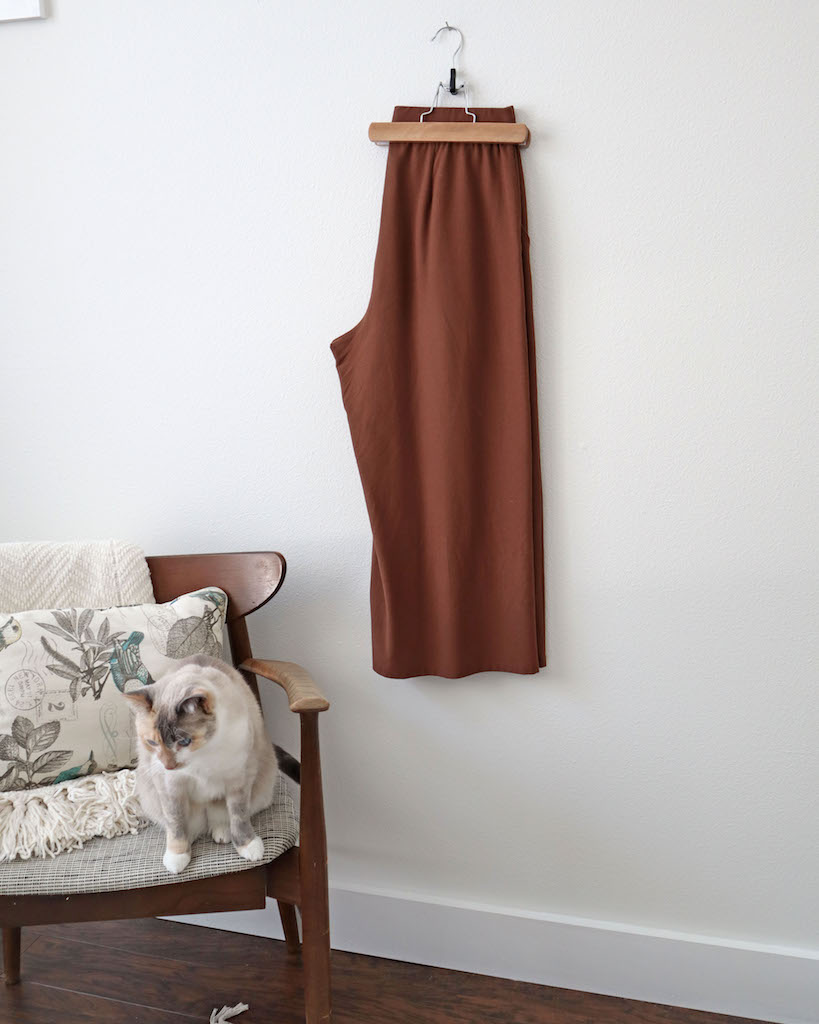 A photo of the fourth item of my winter wardrobe staples which is my brown wide leg pants. They are folded in half and hanging at the waist from a pant hanger. There is a chair off to the side with a cream colored throw and pillow in it. My white and cream cat is sitting in the chair.