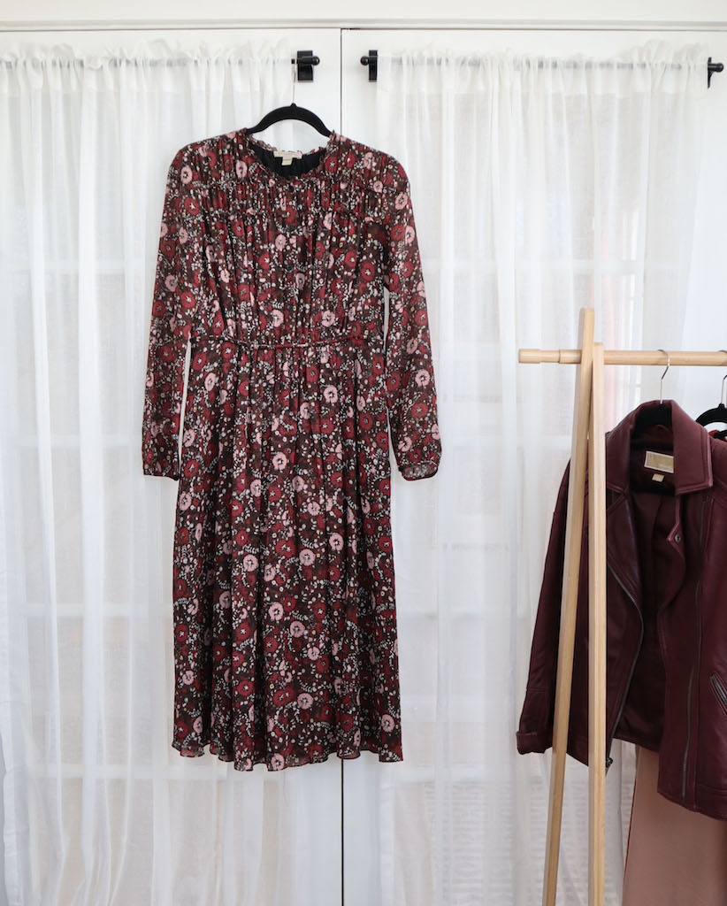 The fifth of my winter wardrobe staples which is my floral dress. It is hanging from a door curtain rail. There is a white sheer curtain behind it. There is a clothing rack off to the right side and you can partially see my Burgundy leather jacket.