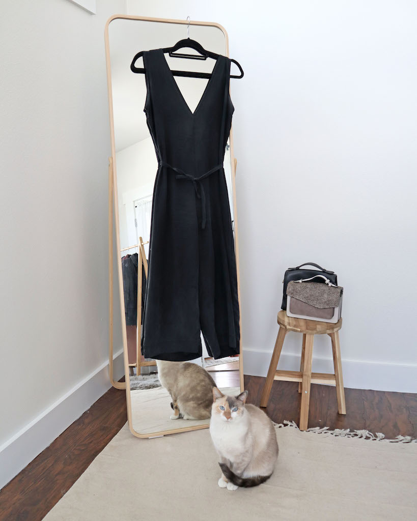 A photo of the second of my winter wardrobe staples which is my black silk jumpsuit hanging over my full length mirror. There is a light wood stool next to the mirror with a grey purse in front of a black purse. My white and grey cat is sitting in fronton the mirror on a cream mat and looking at the camera.