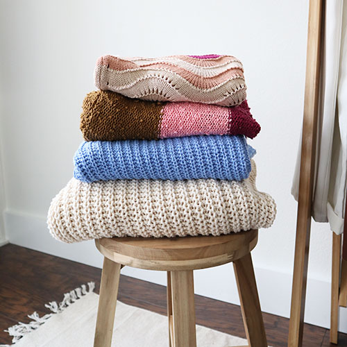 A stack of 4 folded sweaters on a light wood stool that are a part of my cozy capsule wardrobe. On top is a multicolored sweater with light, neutral colors. Under that is a multicolored sweater with more saturated colors. Under that is a cornflower blue sweater on top of a cream colored kind sweater.