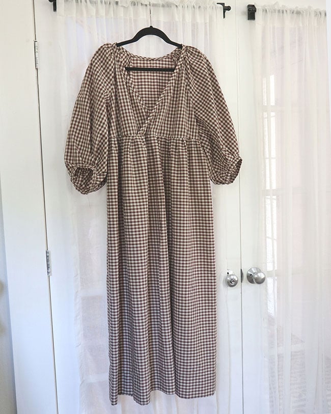 A full length image of a brown and white gingham summer dress that I made for my capsule wardrobe. It is on a hanger and there is a white sheer curtain behind it.