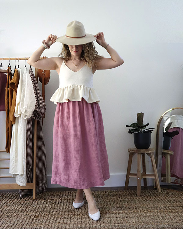 A photo of me wearing the altered white tank top with the ruffled hem over a blush pink skirt with white slingback shoes. I am also wearing a beige wide brimmed hat and holding a tan handbag.
