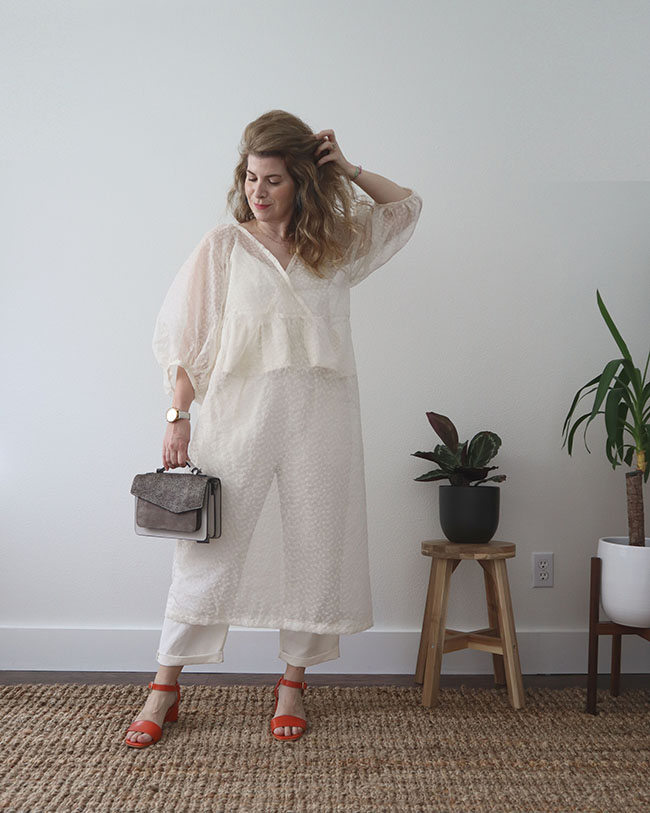 A white sheer long dress with a crossover neckline and puffy sleeves is worn over a white tank top cami with a ruffled hem and white pants. It is paired with orange scrappy sandals and a grey printed bag.