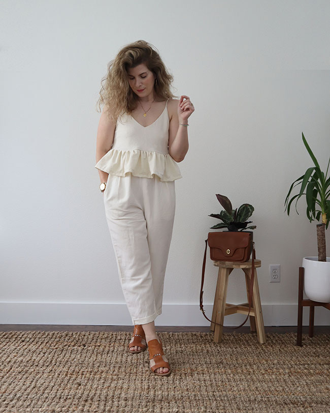 Mini capsule outfit 7 - a white tank top cami with a ruffle hem with a pair of white pleated pants and tan sandals. A tan purse sits on a stool with a. plant.