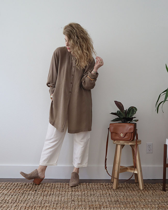Mini capsule outfit 8 - a tan tunic buttoned all the way to the top over white pants with tan leopard print mules. A tan purse sits on a stool with a plant.
