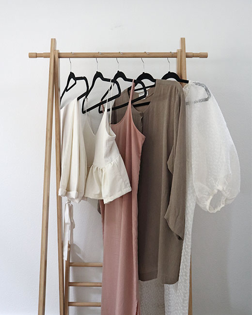The 5 clothing items that I selected for my "capsule by 5" mini capsule wardrobe - from left to right: a pair of white pants, a white tank top with a ruffled hem, a blush pink slip dress, a tan long sleeved tunic, and a white sheer dress with puff sleeves. All items are hanging from a light wood rack.