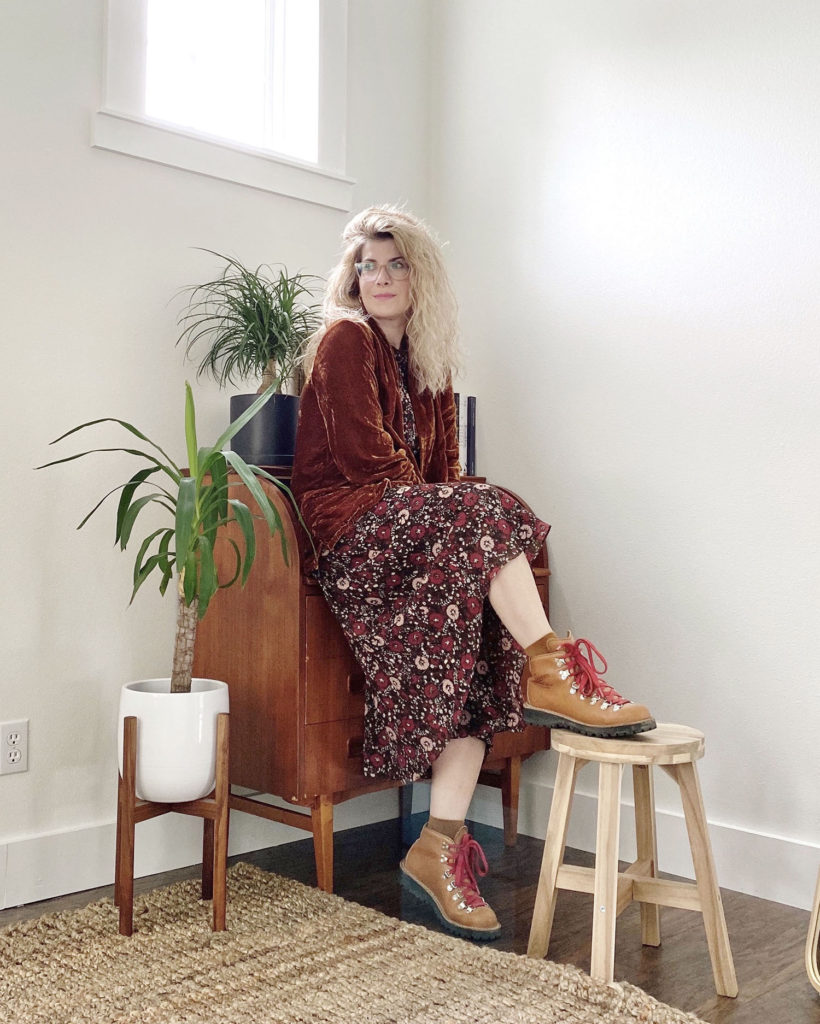 How to style hiking boots in an colorful outfit. A white woman with blonde hair is wearing a trust colored velvet jacket over a dress with pink and red flowers all over and tan hiking boots with red laces. She is sitting on a desk and she has one foot up on a stool.