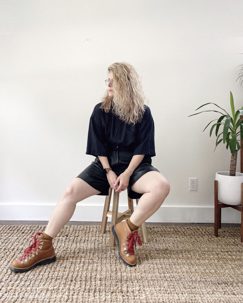 How to style hiking boots in an edgy way. A white woman with blonde hair is sitting on a wooden stool and wearing an oversize black silk blouse tucked into black leather shorts with tan hiking boots with red laces.