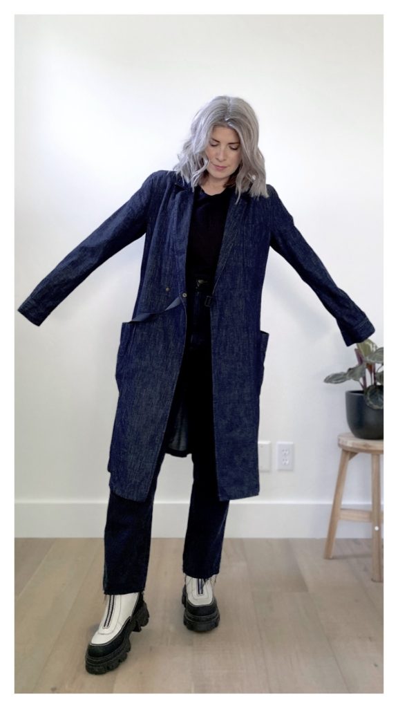 A small white woman with shoulder length silver grey hair is wearing a black t-shirt tucked into black jeans. The is wearing a long dark denim duster that hits just below her knees. She is wearing white platform boots with a thick black sole. She is looking down and has her 2 arms out the the side. Her hands are pulled up into the sleeves.