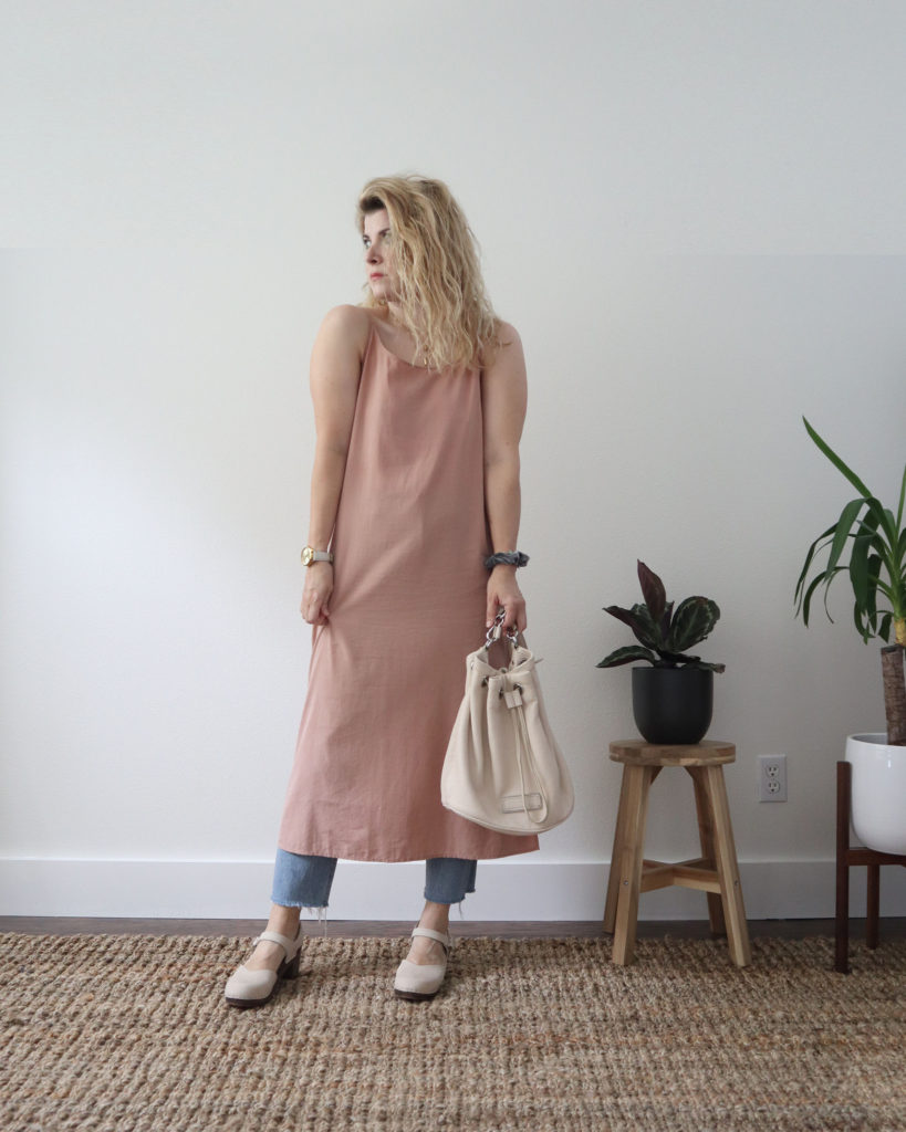 my first dress over pants outfit consists of a blush pink midi length slip dress over a pair of light wash jeans with a cream colored bucket bag and a pair of cream colored clogs with dark wool soles.