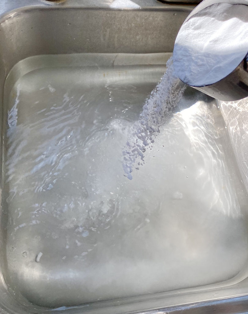 A sink is filled halfway with water and there is baking soda being sprinkled into the water. The baking soda is added to get rid of the mothball smell in the sweater.