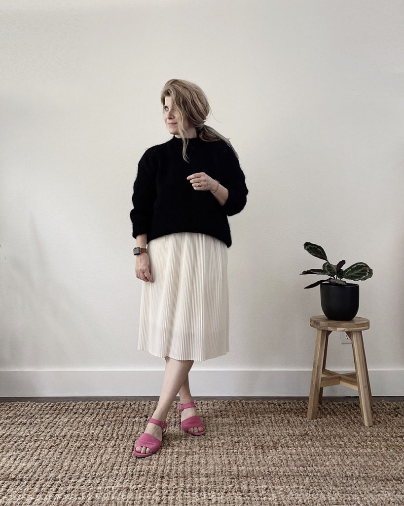 A white woman with blond hair is wearing a black wool sweater after getting the mothball smell out. She is wearing it with a white pleated skirt that is just below the knee with pink suede sandals.