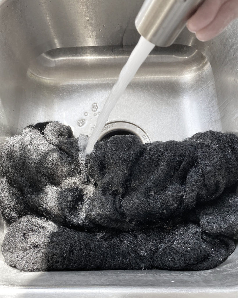 A stream of water from a tap is rinsing the black sweater after the shampoo wash to get rid of any leftover mothball smell.