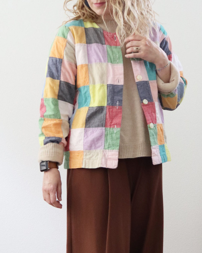 A close up of the colorful patchwork coat. It is worn over an oatmeal sweater and brown pants.