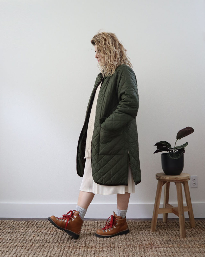 A white woman with wavy blonde hair is wearing a long green quilted coat over a long cream sweater and a cream pleated skirt. She is wearing grey socks and tan hiking boots with red strings. She is standing sideways to the camera.