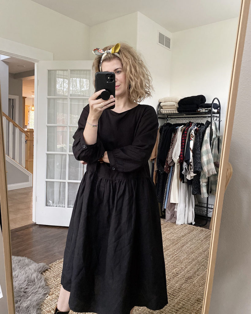A mirror selfie of a blonde woman with short wavy hair. She has her hair pushed back with a bandana that has been made into a headband with the ties on top of her head. She is wearing a loose black long-sleeved midi dress and black combat boots. The outfit remains the same for all of the following pictures.