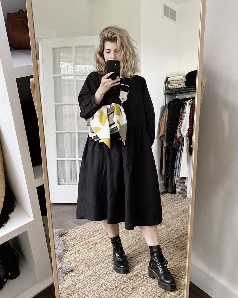 A mirror selfie of a blonde woman with short wavy hair. She is wearing a loose black long-sleeved midi dress and black combat boots. She is also wearing a grey crossbody handbag that she has tied the bandana around.