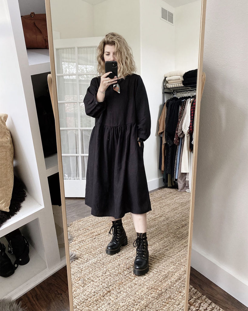 A mirror selfie of a white woman with blond wavy hair who is wearing a long sleeved black dress that is slightly oversized. The dress is midi length and she is wearing black combat boots.