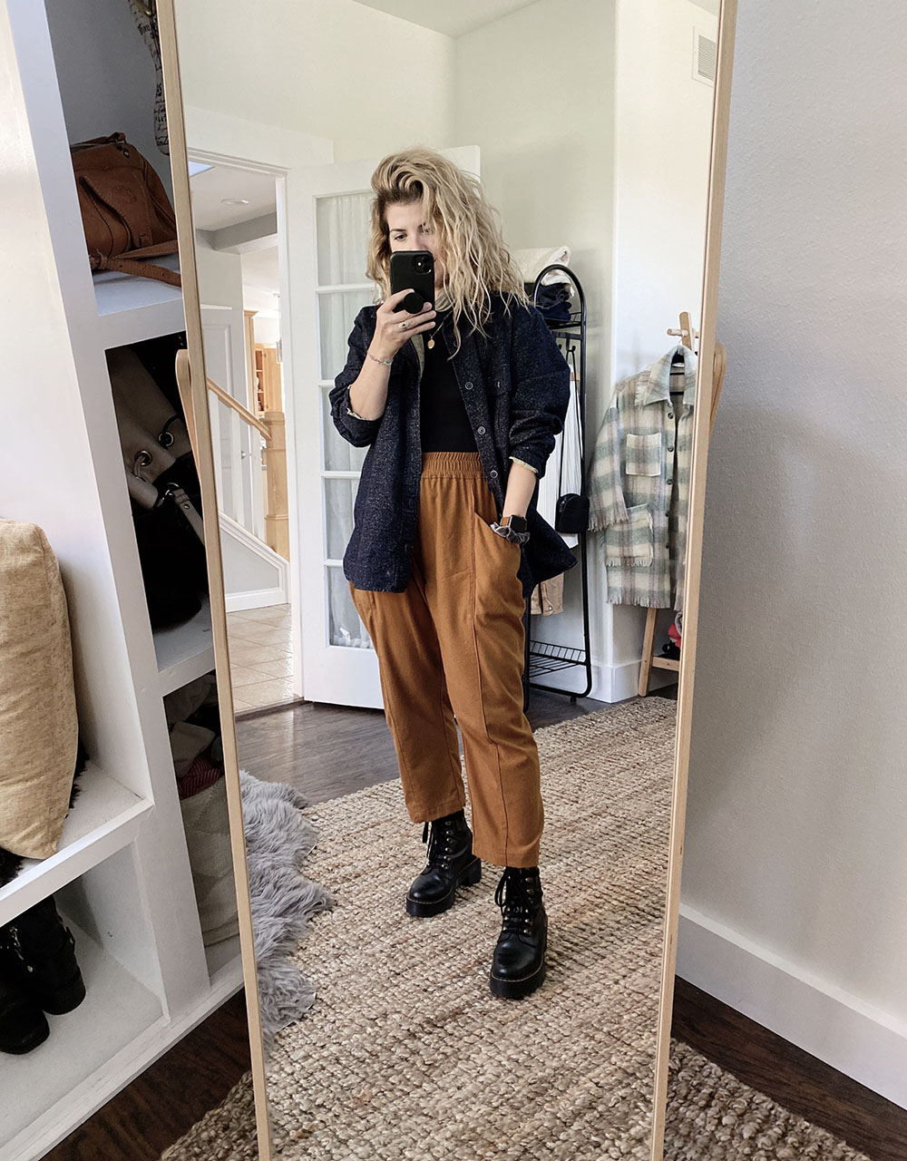 A white woman with blonde wavy hair is wearing a navy blue shirt unbuttoned over a black top and rust colored pants. She is also wearing black combat boots.
