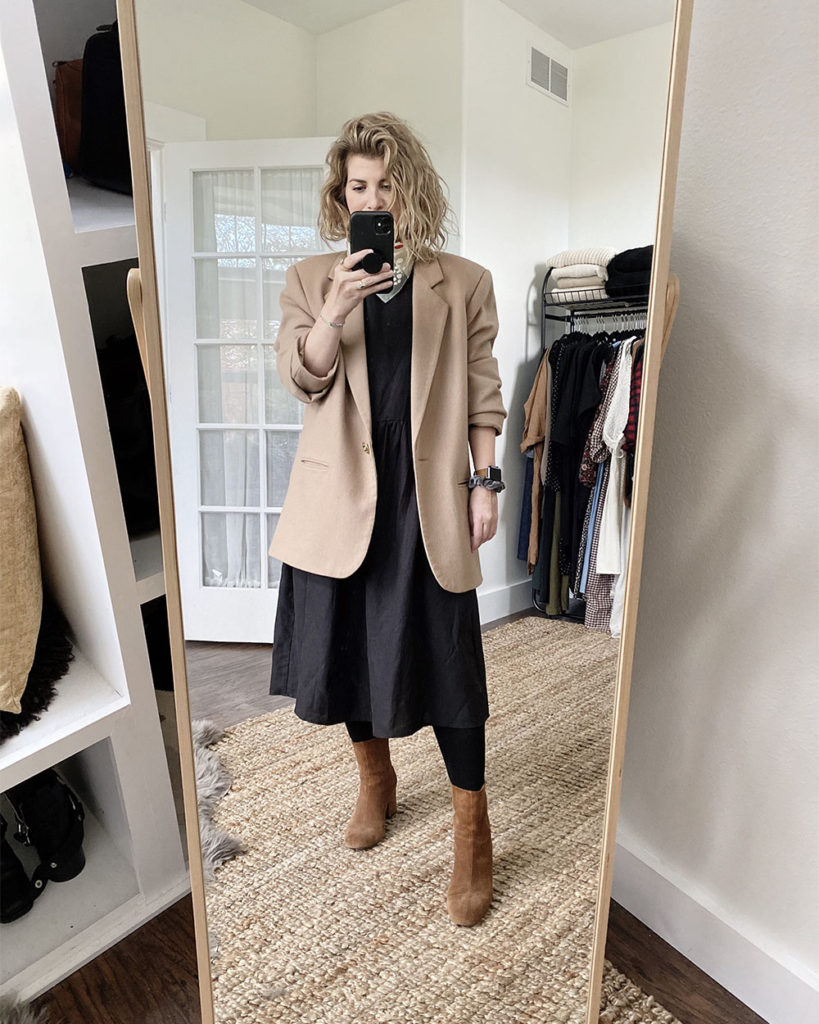 A mirror selfie of a white woman with blond wavy hair who is wearing a long sleeved black dress under an oversized tan blazer. She has a bandana around her neck and she is wearing black tights and brown boots.