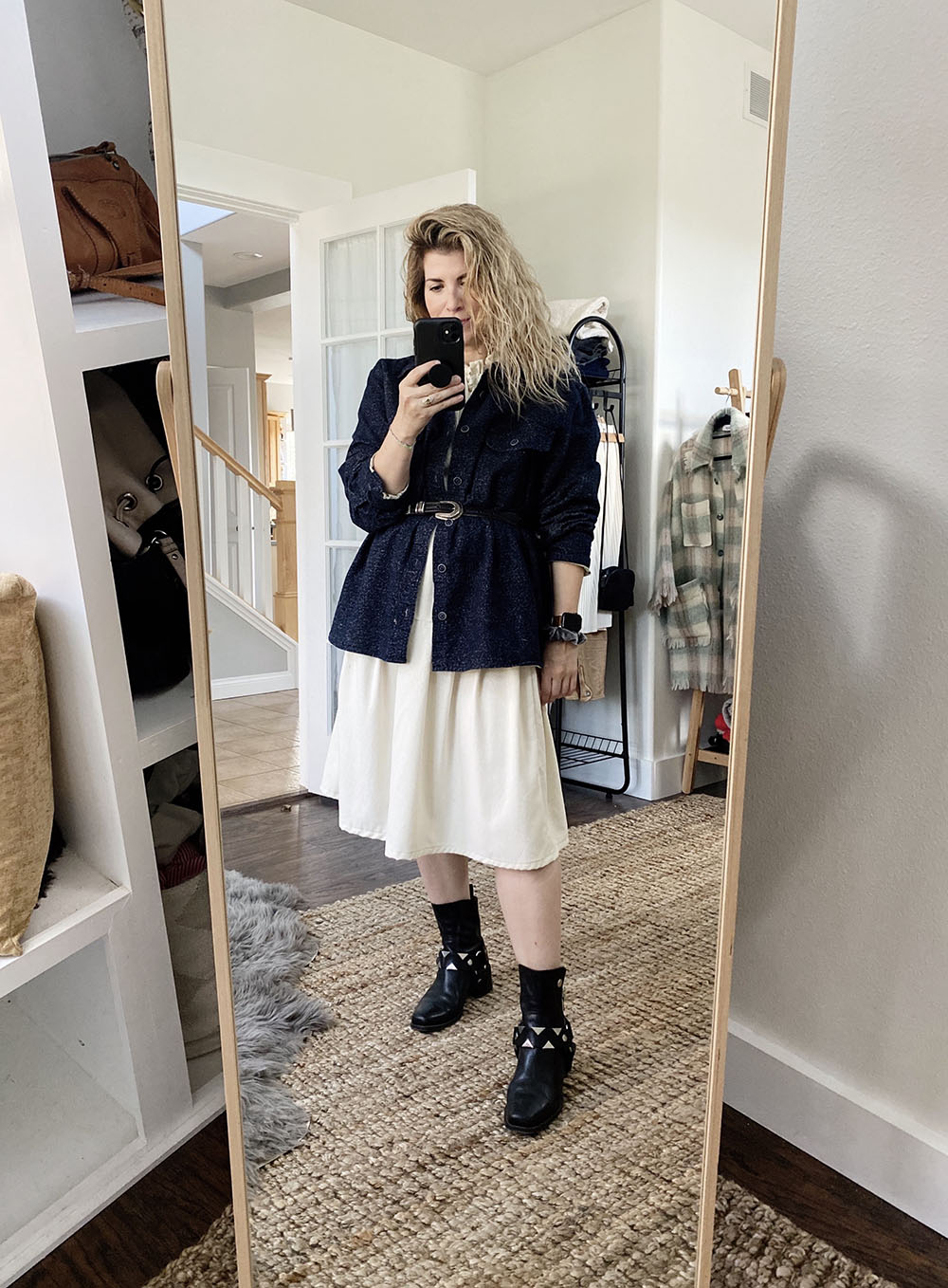 A white woman with blonde wavy hair is wearing a navy blue shirt unbuttoned over a cream colored dress. She has the shirt and dress belted at the waist with a black belt with a silver buckle. She is wearing black moto boots with silver arrow detailing.