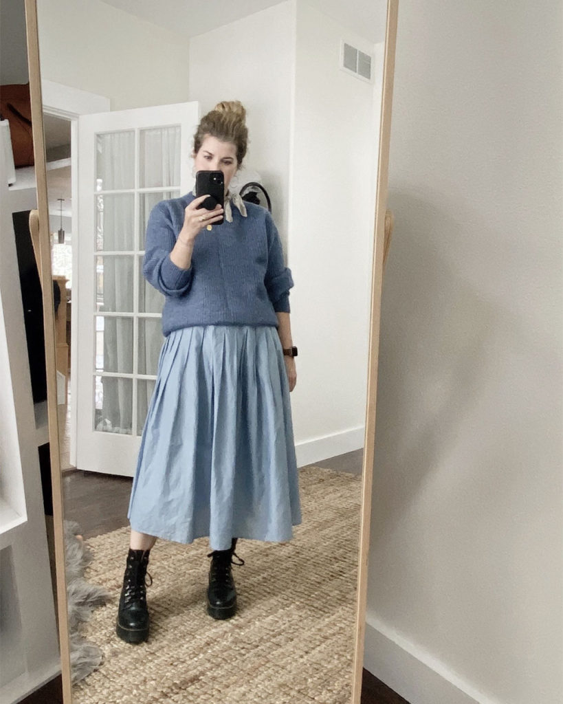 Sweater Outfit 2 - a mirror selfie of a white woman with wavy blonde hair wearing a blue crew neck sweater over a blue pleated waist dress. She has a grey scarf tied around he neck and is wearing black combat boots. The next 3 photos are closeups or different angles.