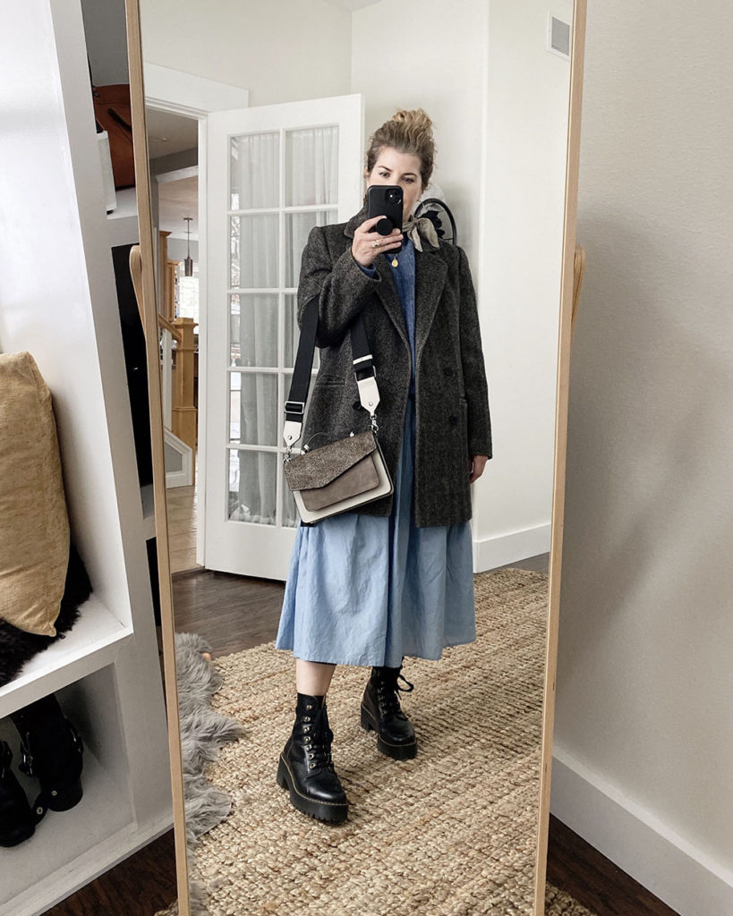 Sweater Outfit 2 - a mirror selfie of a white woman with wavy blonde hair wearing a blue crew neck sweater over a blue pleated waist dress. She has a grey scarf tied around he neck and is wearing black combat boots. She is wearing a grey overcoat and has a great handbag over her arm.