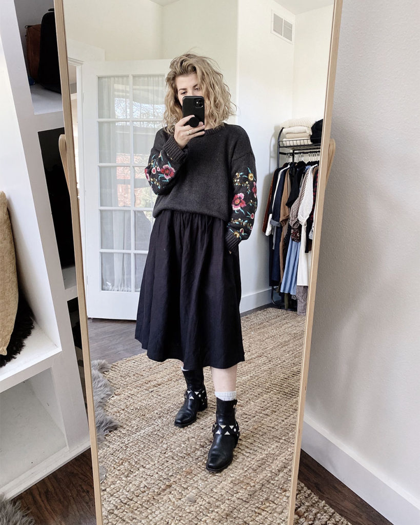 A mirror selfie of a white woman with blond wavy hair who is wearing a grey sweater with floral embroidery on the sleeves over a long sleeved black dress that is slightly oversized. The dress is midi length and she is wearing black moto boots with silver detailing.